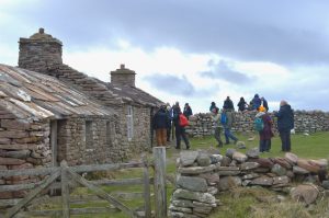 Photo of people in winter clothing walking around a stone-built bothy with dystone walls in the foreground
