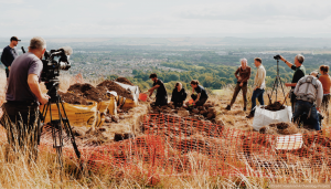 A large group of people surrounding an archaeological trench while filming takes place