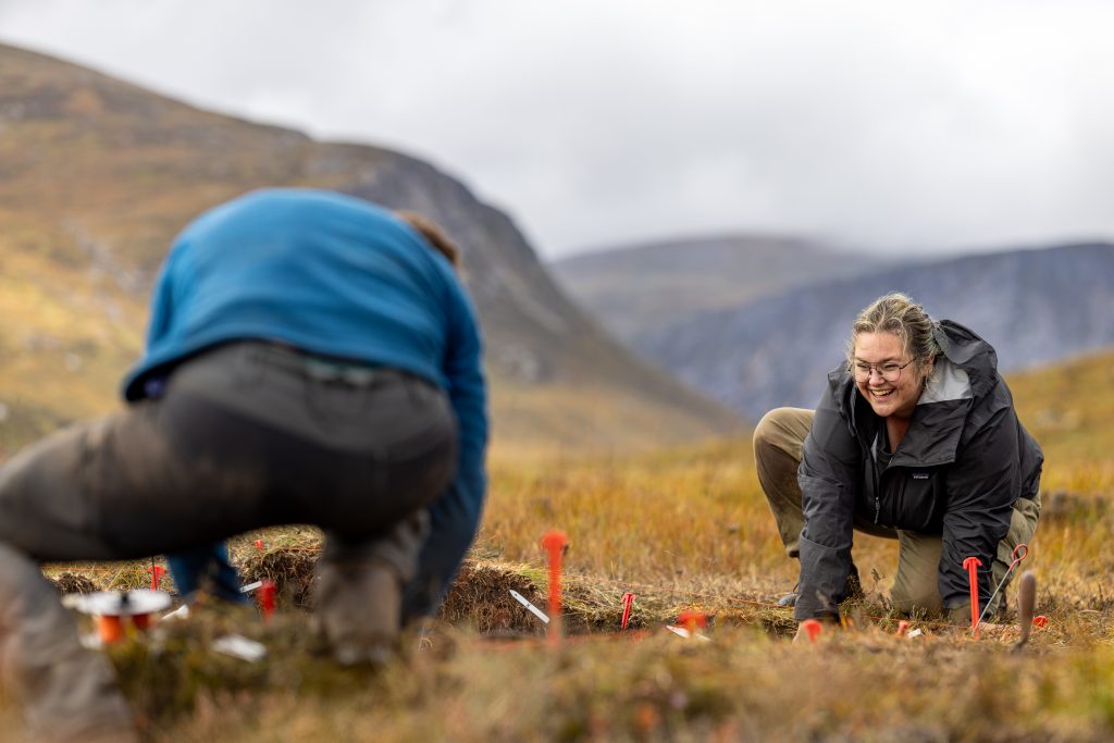 Two people crouching near an archaeological trench in a mountainous setting with one person (a student) smiling at the other