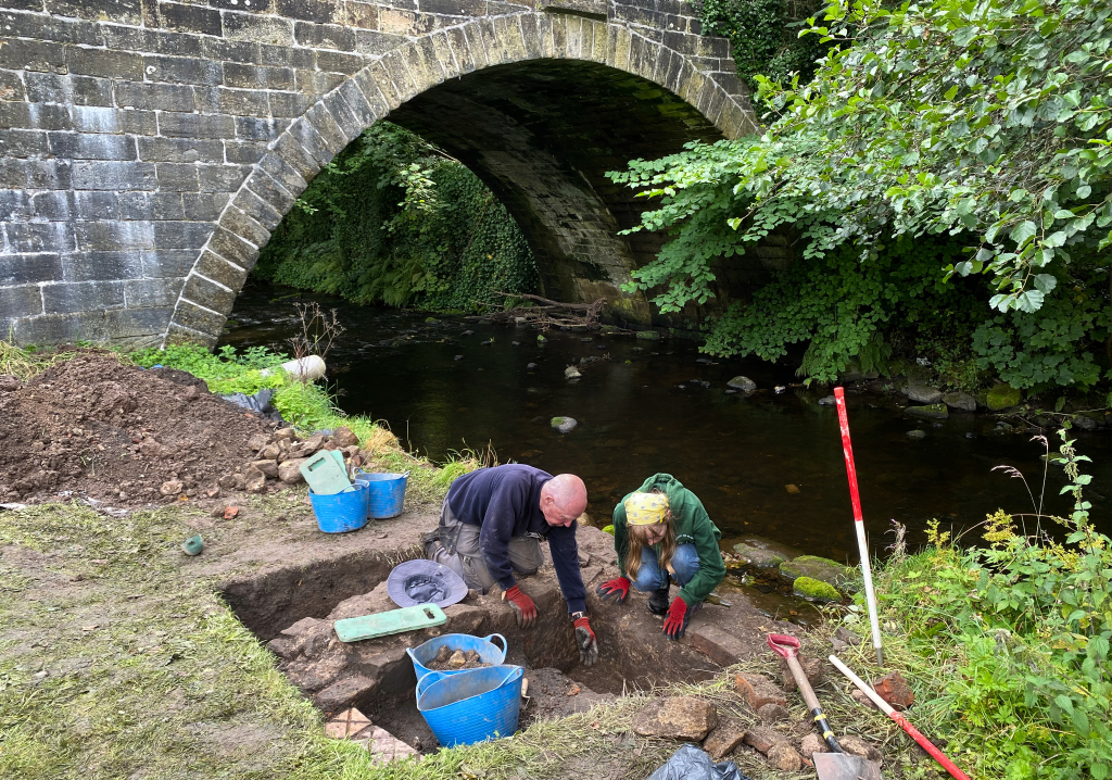 Two people surrounded by archaeological tools , such as shovels and buckets , digging in a trench beside a river with an old stone bridge in the background