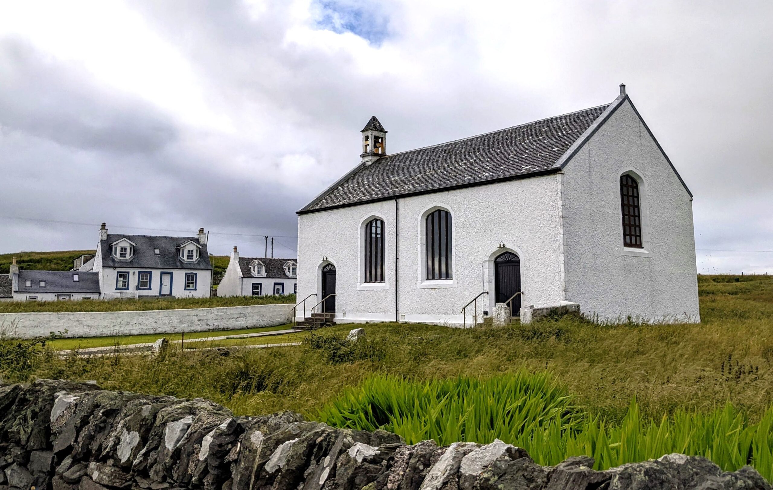 Photo of a white rectangular plan church in a green field with a dry stone wall in the foreground