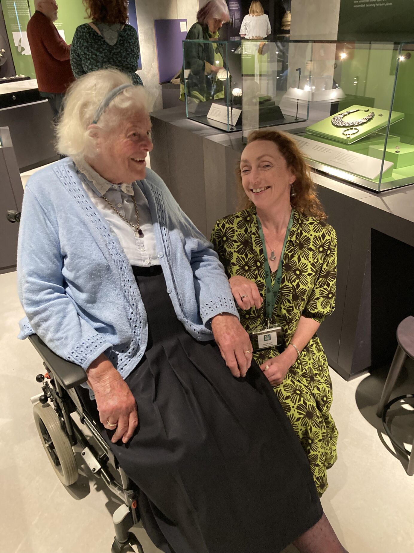 Photo of an elderly person in a wheelchair and a person in a green dress sitting next to her