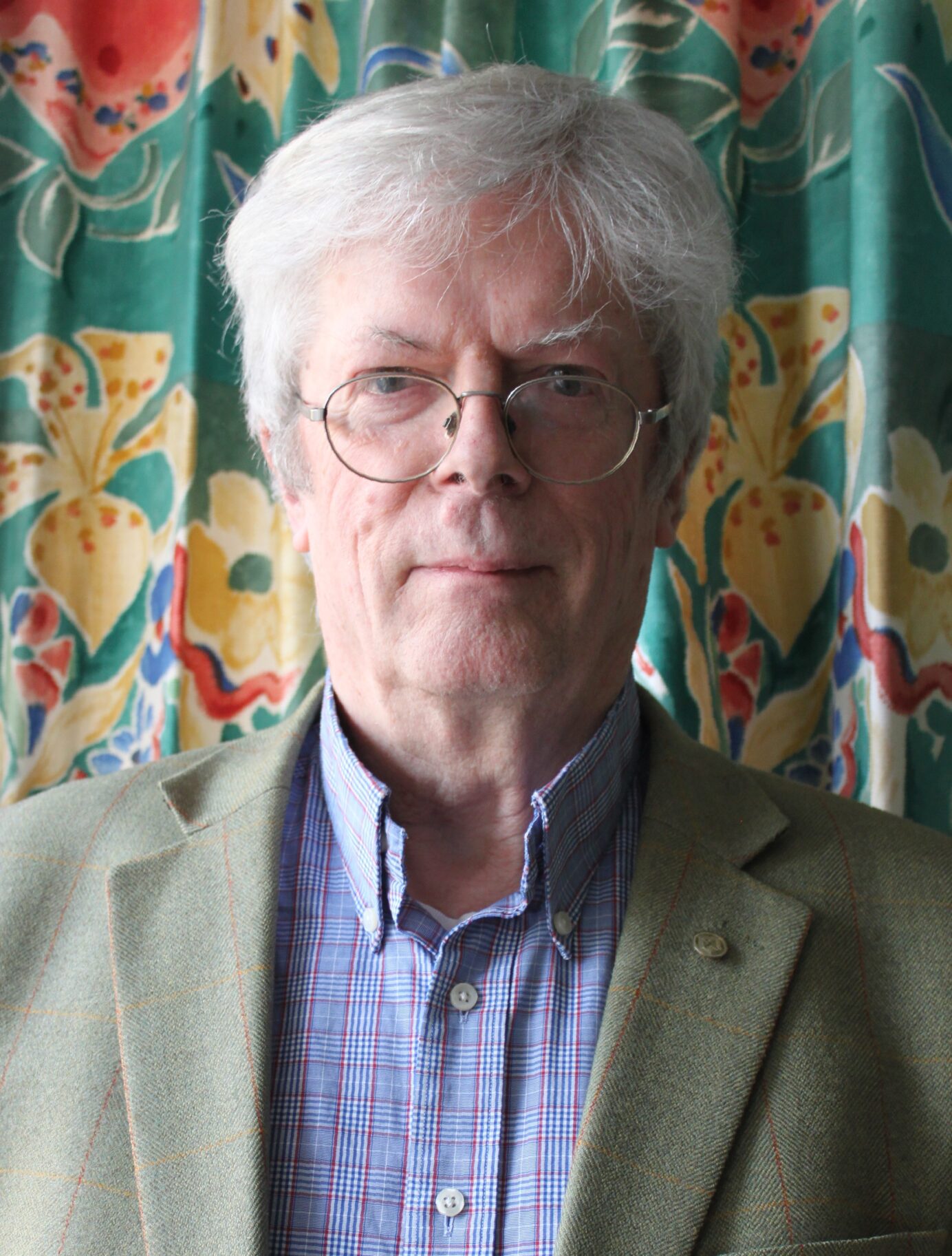 Photo of a man with short white hair, glasses wearing a blue check shirt and green blazer