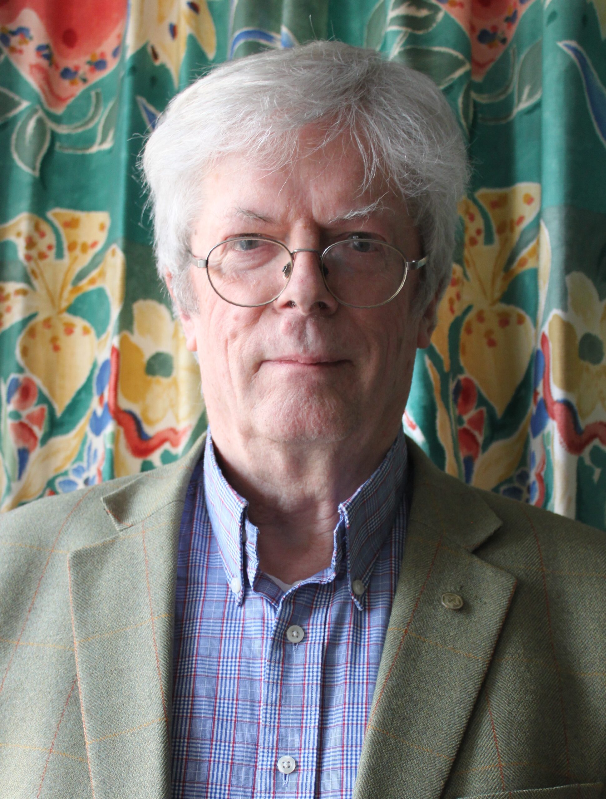 Photo of a man with short white hair, glasses wearing a blue check shirt and green blazer