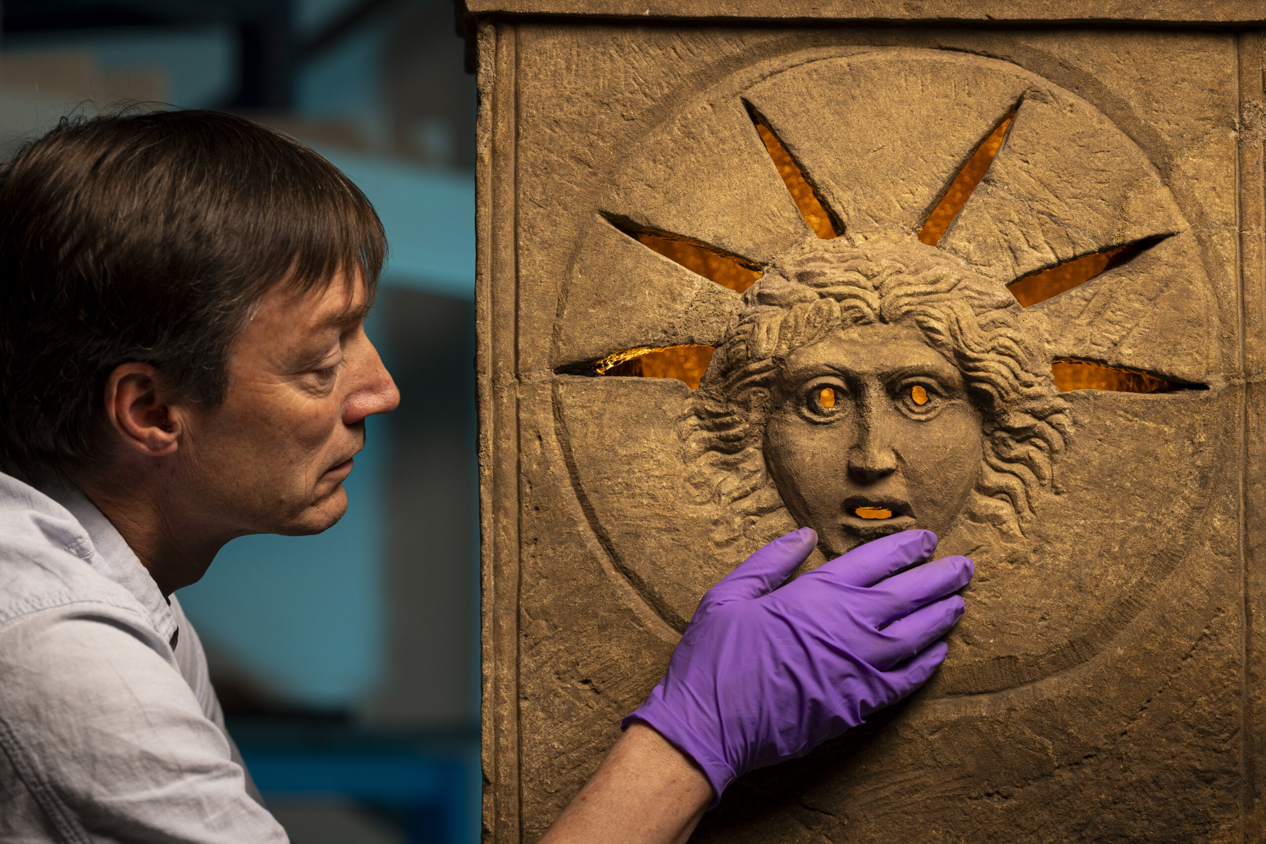 Curator Dr Fraser Hunter examines the Roman altars.Photo © Duncan McGlynn (4) A person wearing purple gloves touching a Roman altar which depicts the face of the sun god Sol and would have been illuminated from behind so that in the low light of the temple, worshippers would have seen his eyes, mouth and the rays of his solar crown glowing.