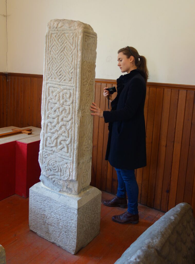Photo of a person with brown hair tied in a ponytail wearing a long black coat and holding a digital camera, standing inside a small building and looking at an incised stone slab